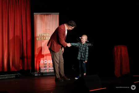 Magik wita na scenie chłopczyka w koszuli w kratkę. Chłopczyk podaje rękę magikowi.
