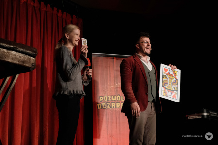 Dziewczyna w szarym swetrze pokazuje małą kartę, taką samą jaką trzyma Magik. Dziewczyna śmieje się.