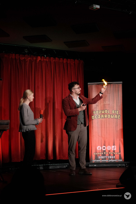 Z dłoni magika wychodzi mały żółty płomień. Dziewczyna w szarym swetrze patrzy się i uśmiecha.