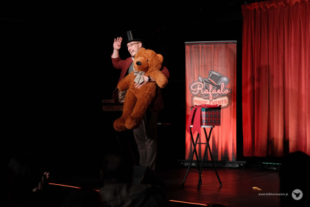 Magik ma założony czarny cylinder. Trzyma wielkiego brązowego misia.