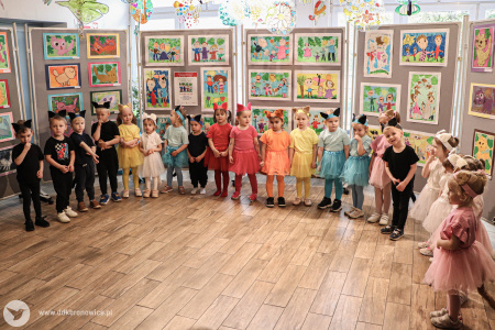 Galeria Mała. Dzieci stoją w półkolu. Chłopcy ubrani na czarno. Dziewczynki w kolorowe spódniczki. Za nimi wiszą obrazy.