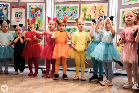 Dziewczynki w kolorowych ubrankach tańczą i śmieja się. Za nimi widać powieszone obrazy małych artystów.