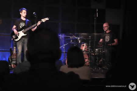 Na scenie po lewej stronie stoi gitarzysta. Trzyma czarno-białą gitarę basową. Po prawej stronie muzyk gra na perkusji.