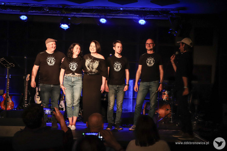 Zespół TB Squad stoi razem na scenie. Mają czarne-białe koszulki zespołu. W skład zespołu wchodzą dwie kobiety i trzech mężczyzn.