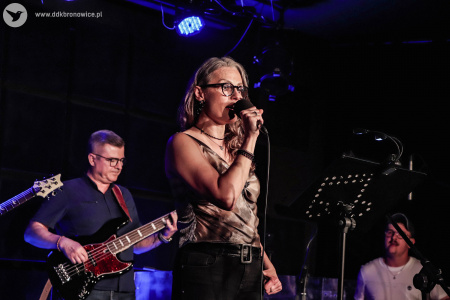 Wokalistka śpiewa na scenie do mikrofonu. W tle widać dwóch gitarzystów i po prawej stronie perkusista.