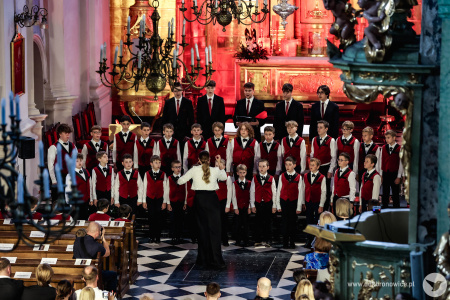 Irena Sołtan-Wąsik dyryguje chórem chłopięcym ubranym w czerwone kamizelki. W tle delikatne czerwone światło podkreśla złote ornamenty kościoła. Widać siedząca publiczność.