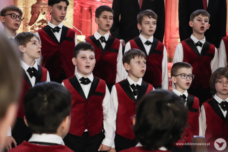 Irena Sołtan-Wąsik dyryguje chórem chłopięcym ubranym w czerwone kamizelki. W tle delikatne czerwone światło podkreśla złote ornamenty kościoła.