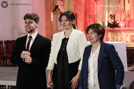 W środku, w białym żakiecie, stoi Irena Sołtan-Wąsik, dyrygentka Chóru Chłopięcego Słowiki Lubelskie. Po jej prawej mężczyzna w czarnym garniturze, który pokazuje sygnet na palcu. Po lewej stronie kobieta w granatowym kostiumie.