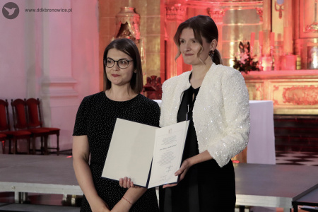 Irena Sołtan-Wąsik pozuje z oficjalnym dyplomem. Obok niej stoi kobieta w czarnej sukience w białe drobne kropki.