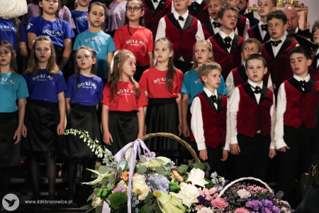Występ trzech zespołów. Chóru Dziewczęcego DO-RE-MI, Chóru Chłopięcego Słowiki Lubelskie i Męskiego Zespołu KAIROS. Za nimi czerwone światło oświetla złote fragmenty i białe ściany kościoła. Przed zespołami widać postawione kwiaty.
