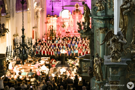 Występ trzech zespołów. Chóru Dziewczęcego DO-RE-MI, Chóru Chłopięcego Słowiki Lubelskie i Męskiego Zespołu KAIROS. Za nimi czerwono-fioletowe światło oświetla złote fragmenty i białe ściany kościoła.