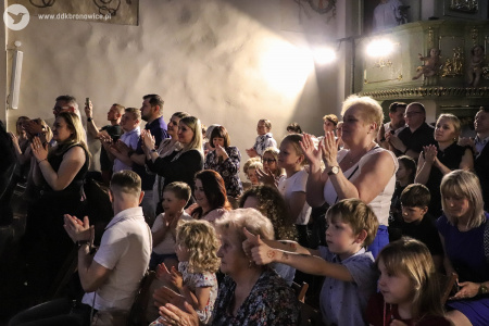 Publiczność wstała z miejsc. Klaszczą w dłonie. Unosi się lekko biały dym. Przez dym przebija się światło z reflektora.