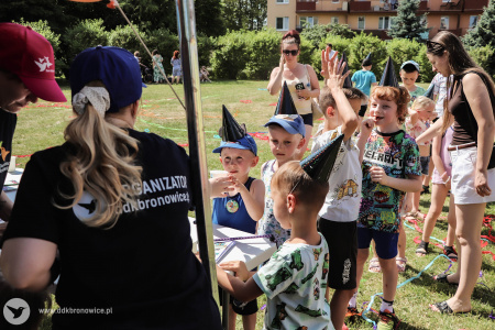 Po lewej stronie widać osobę z czarną koszulką. Na koszulce napis: ORGANIZATOR. DDK BRONOWICE. Ma założoną granatową czapkę. W kolejce do tej osoby ustawione są dzieci.