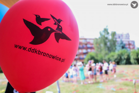 Czerwony balon. Na nim czarny napis: WWW.DDKBRONOWICE. Obok napisu trzy czarne kolibry. W tle widać zabawy wokół namioty klaunów.