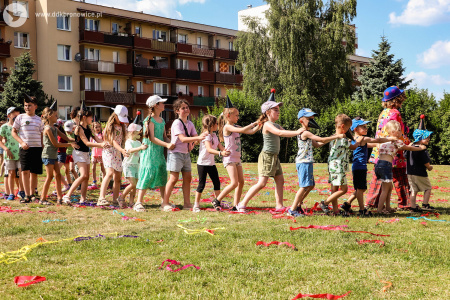 Dzieci trzymają się za ramiona. Idą gęsiego. Dzieci prowadzi na przodzie klaun w niebieskim meloniku.