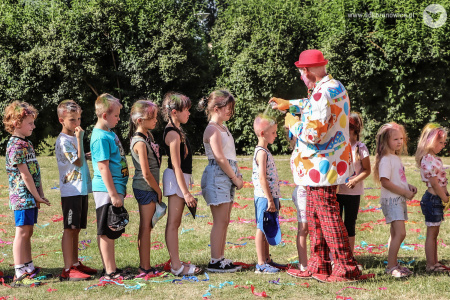 Klaun w czerwonym meloniku koloruje włosy dzieciom. Dzieci ustawione są w rzędzie.