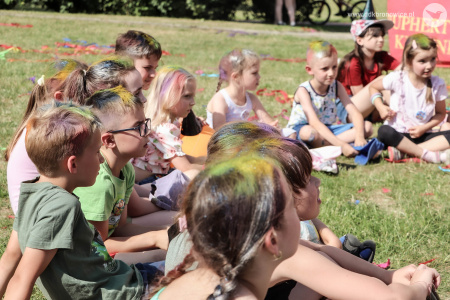 Grupa dzieci siedzi na trawie. Niektóre z nich maja pomalowane włosy na kolorowo.