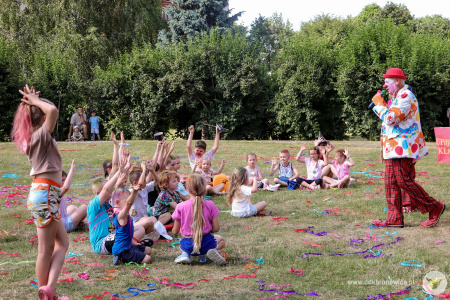 Klaun w czerwonym meloniku rozmawia z dziećmi. Dzieci wysoko podnoszą rękę jakby się zgłaszały do odpowiedzi.