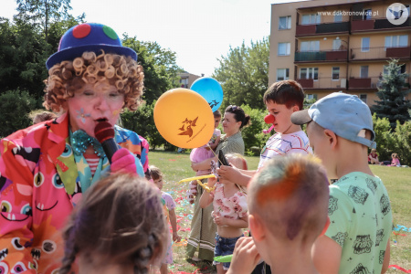 Klaun w niebieskim meloniku mówi do mikrofonu i podchodzi do grupki dzieci.
