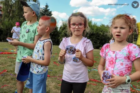Czwórka dzieci stoi i przygląda się zabawom. W rękach trzymają naklejki rozdane przez klaunów.