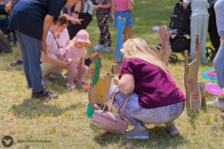 Z ziemi wystaje figurka tekturowego koguta. Mama trzyma na rękach dziecko, które maluje zwierzę.