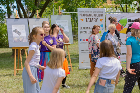 Animatorki prowadzą zabawy ruchowe dla dzieci. Wokół nich zgromadzona grupa dzieci. W tle widać rollupy reklamujące DDK Bronowice.