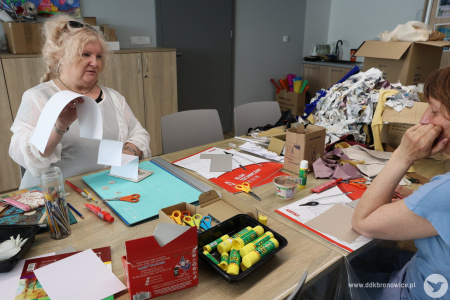 Prowadząca warsztaty Katarzyna Kate Bauer tłumaczy jak wykonać książkę harmonijkową. Na stole kartki, kleje, nożyczki i przedmioty papiernicze.