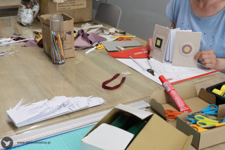 Na stole kartki, kleje, nożyczki i przedmioty papiernicze. Uczestniczka w niebieskiej koszulce  tworzy swoja książkę harmonijkową.