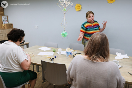 Pracownie Kultury Dziesiąta. Uczestniczki tworzą pająki ludowe przy jasnym stole warsztatowym. Emilia Skubis pomaga uczestniczkom. Z sufitu zwisa zielony pająk.