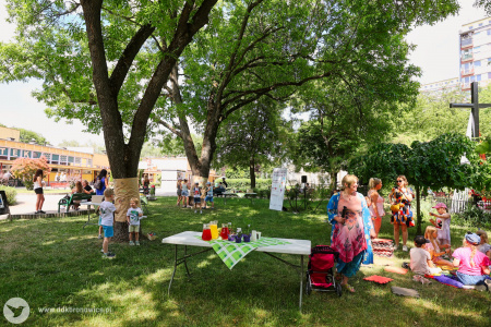 Piknik pod drzewami. Po prawej stronie siedzą dzieci i bawią się na trawie. Widać stół z sokami i owocami.