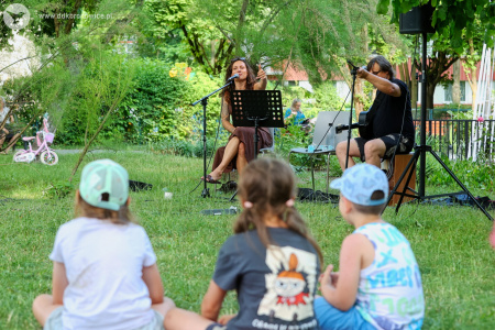Trójka dzieci słucha koncertu. W tle Sylvetta i gitarzysta grają koncert. Siedzą na jasnych krzesłach. Ustawione mikrofony. Artystka gra na tamburynie i śpiewa.
