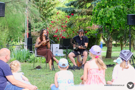 Dzieci słuchają uważnie słuchają koncertu. Trójka dzieci słucha koncertu. W tle Sylvetta i gitarzysta grają koncert. Siedzą na jasnych krzesłach. Ustawione mikrofony. Artystka gra na tamburynie i śpiewa.