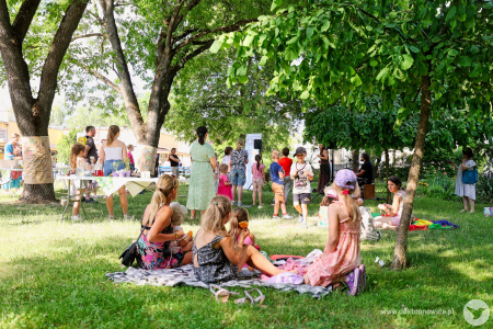 Kobiety i dzieci leża na kocach. Słuchają koncertu. Obok stół z sokami i owocami.