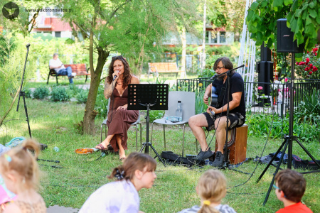 Sylvetta w brązowej sukience śpiewa piosenkę. Trzyma mikrofon. Obok niej gra gitarzysta w czarnej koszuli.