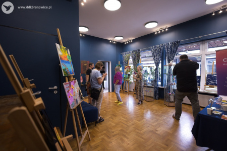 Galeria w pasażu. Widac zawieszone obrazy, obrazy na sztalugach, ozdoby w gablotach. Ludzie oglądają wystawę. Mężczyzna w czarnej koszuli robi zdjęcia aparatem.