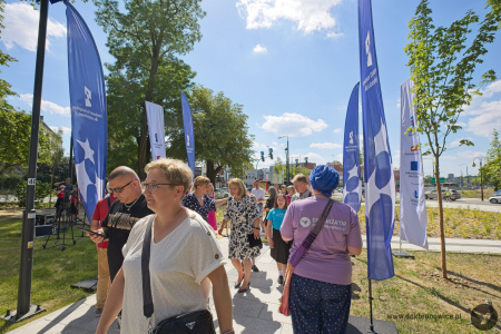 Mieszkańcy Lublina przychodzą na otwarcie parku. Idą chodnikiem. Widać niebieskie flagi Funduszy Europejskich i wysokie drzewa.