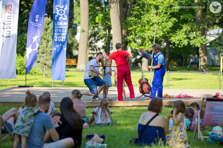 Aktorzy rozmawiają na scenie z zaproszonymi dziećmi. Przed drewnianą sceną na trawie siedzi publiczność.