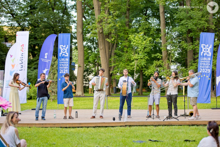 Orkiestra występuje na drewnianej scenie. Są skrzypce, trąbki, puzony, bęben, akordeon.