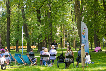 Muzyk w ciemnych okularach gra na gitarze do mikrofonu. Przed nim, na leżakach siedzi publiczność.