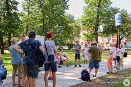 Spacer po parku z przewodnikiem. Grupa ludzi uważnie słucha ciekawostek. Jest bardzo słonecznie. Widać drzewa i infrastrukturę poza parkiem. Widać duże zielone drzewa. em.