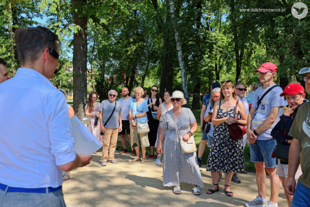 Spacer po parku z przewodnikiem. Grupa ludzi uważnie słucha ciekawostek. Jest bardzo słonecznie. Widać drzewa i infrastrukturę poza parkiem. Widać duże zielone drzewa.