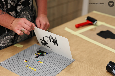 Instruktorka odbija pomalowane klocki lego na białą kartę. Na kartce powstaje charakterystyczny czarny wzór.