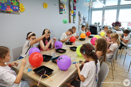 Dzieci tworzą ozdoby do nadmuchanych kolorowych balonów. Na balonach widać napis: www.ddkbronowice.pl i trzy czarne kolibry.