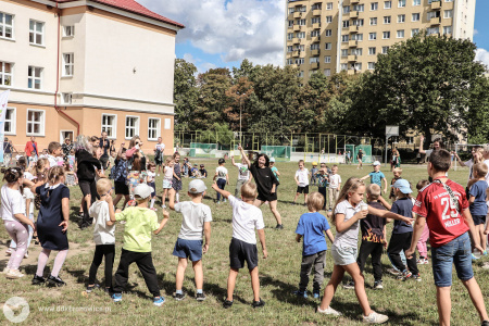 Na boisku dzieci robią rozgrzewkę ruchową. Stoją w kółku, w środku dziewczynka prowadzi ćwiczenie, które pozostałe dzieci muszą wykonywać.