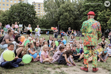Klaun w czerwonym meloniku i zielonym garniturze rozmawia z dziećmi przez mikrofon. Dzieci są uśmiechnięte.