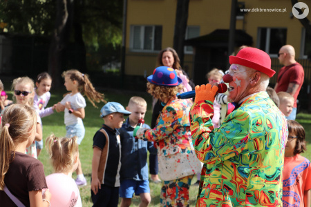 Klaun w czerwonym meloniku i zielonym garniturze rozmawia z dziećmi przez mikrofon. Dzieci są uśmiechnięte.