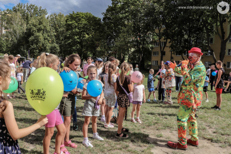 Klaun w czerwonym meloniku i zielonym garniturze rozmawia z dziećmi przez mikrofon. Dzieci są uśmiechnięte.