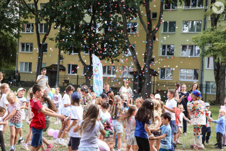 Dzieci tańczą w parach. Obok nich bawi się klaun w niebieskim meloniku. W powietrzu unosi się wystrzelone confetti.