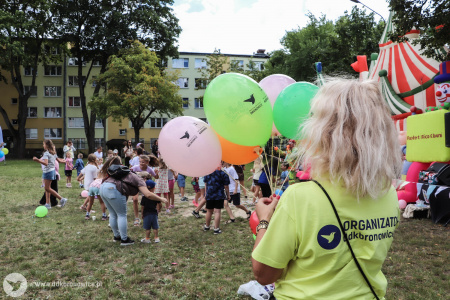 Pracowniczka Dzielnicowego Domu Kultury Bronowice ma założoną żółtą koszulkę firmową. Trzyma pęk nadmuchanych balonów. W tle klauni bawią się z dziećmi.
