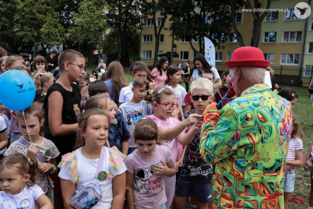 Klaun w czerwonym meloniku rozmawia z dziećmi przez mikrofon. Przed nim stoi duża grupa dzieci.